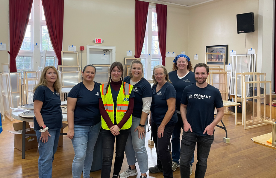 Versant Power team members gathered at a community event location featuring wooden structures and red curtains in the background.
